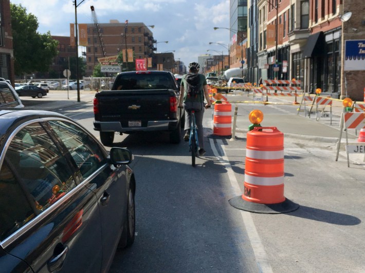 A person rides a Divvy bike north on Milwaukee Avenue and squeezes between cars and orange traffic barrels.