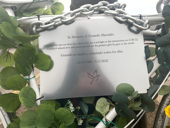 Close up of a metal placard affixed to a white ghost bike. A close-up of the placard hanging on the ghost bike in remembrance of Gerardo Marciales. It reads: "In memory of Gerardo Marciales: Gerardo‘s life was cut short by a driver who ran a red right at this intersection Gerardo sincere love and passionate life are the greatest gift he gave to the world. Gerardo es amado y extrañado todos los días. (Gerardo is loved and missed every day) 10/9/1980-3/2/2022"