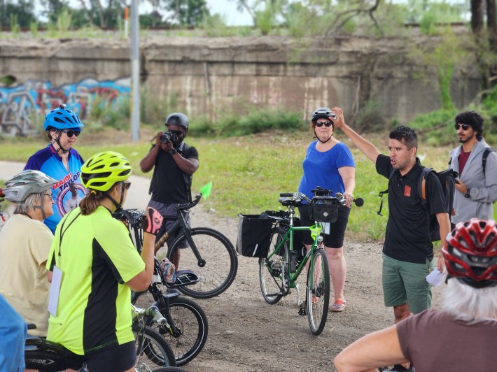 Tour leader Alan Oviedo discusses bike access challenges with a group of bikers, between the expressway and train viaducts.