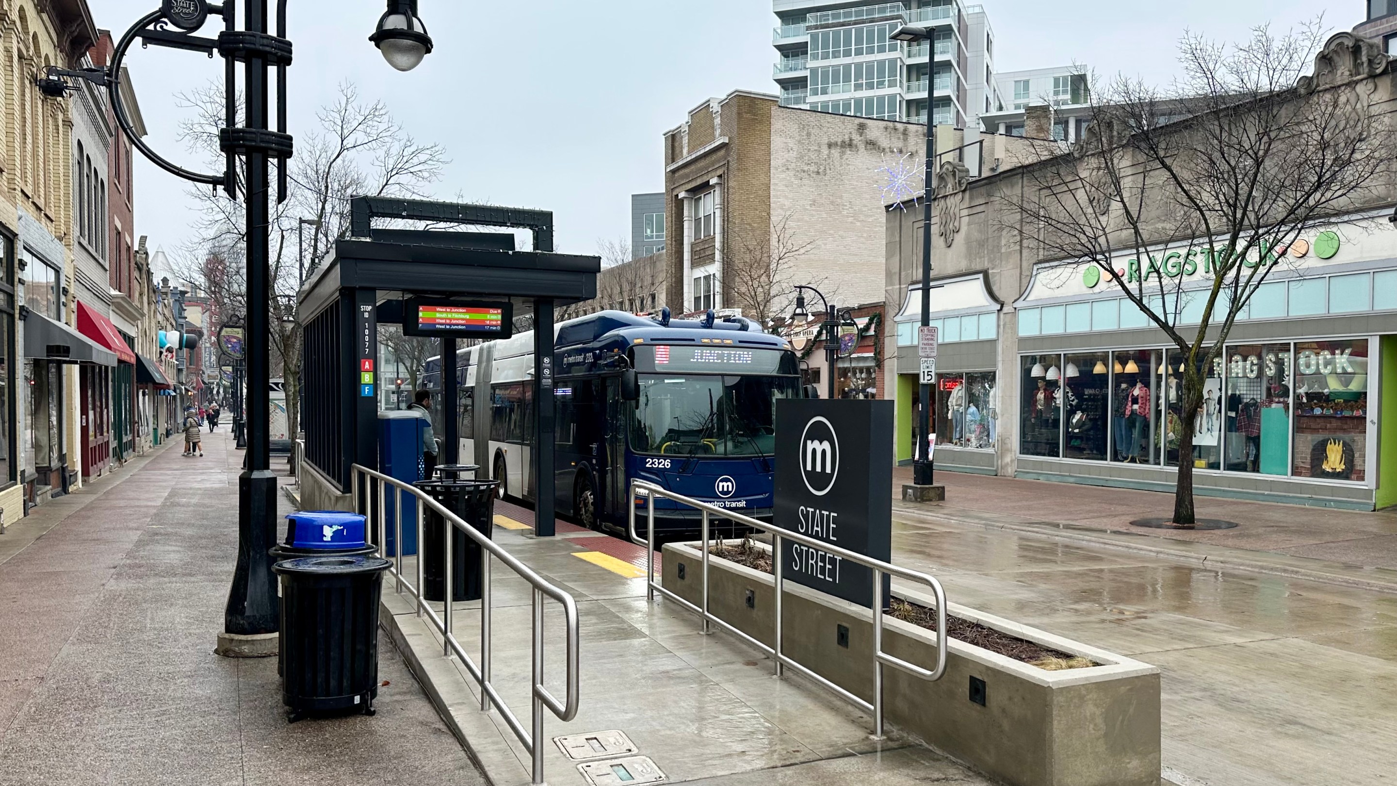 photo of an articulated bus docking at the State Street station in Madison, Wisconsin. The street is pedestrianized and has a lot of local and national stores and restaurants.