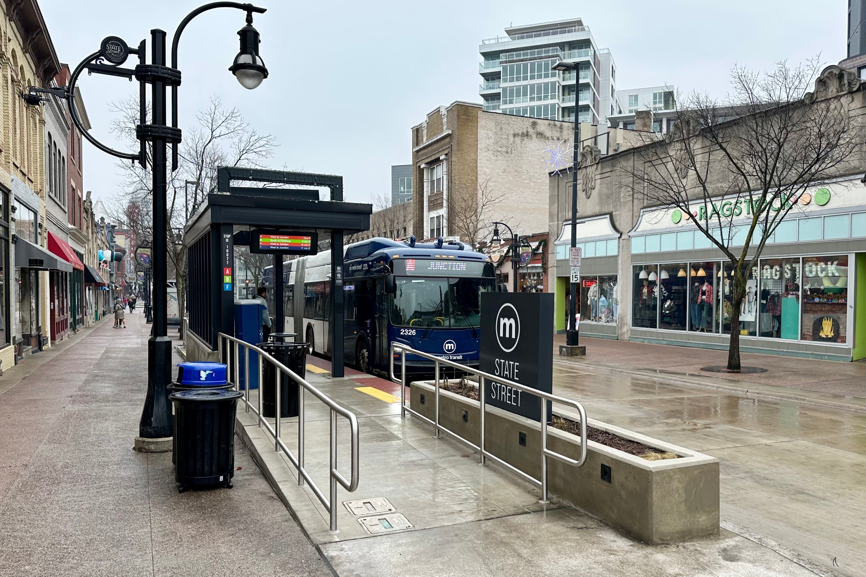 photo of an articulated bus docking at the State Street station in Madison, Wisconsin. The street is pedestrianized and has a lot of local and national stores and restaurants.