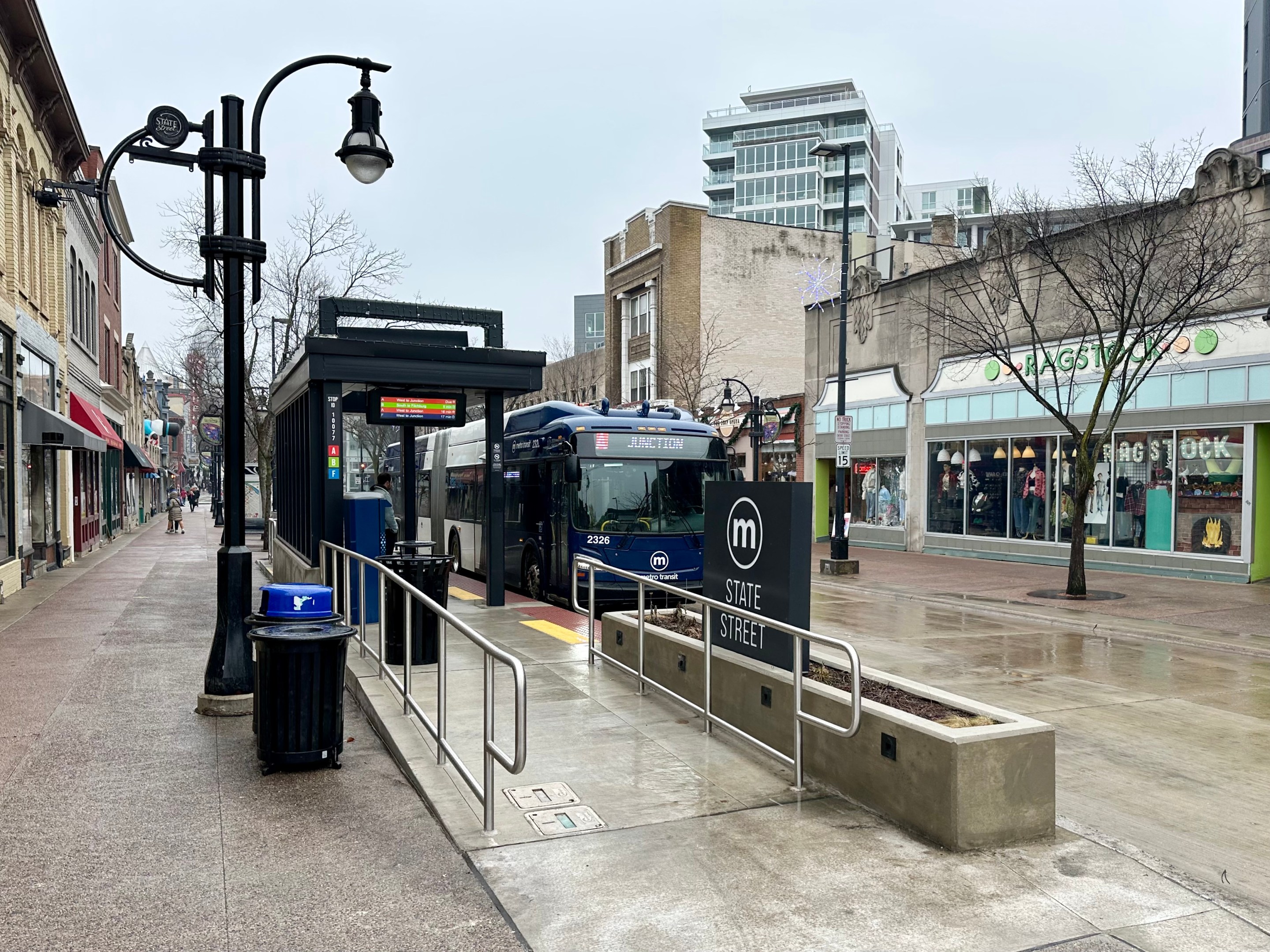 photo of an articulated bus docking at the State Street station in Madison, Wisconsin. The street is pedestrianized and has a lot of local and national stores and restaurants.